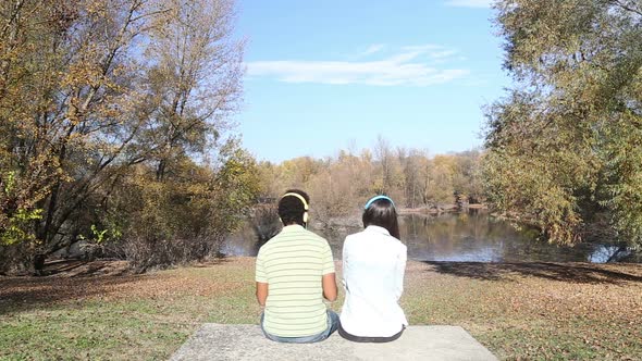 Man And Woman Listening To Music Using Headphones While Sitting By Lake 1 alt