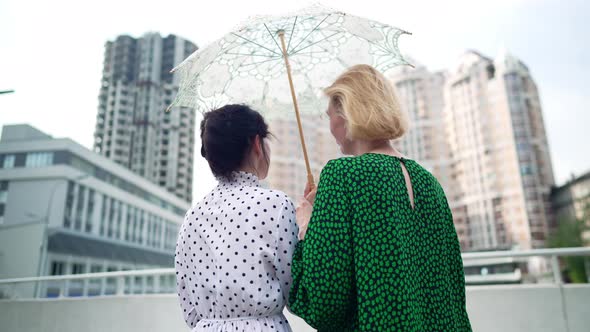 Back View Two Happy Women in Elegant Dresses with Sun Umbrella Standing Outdoors Admiring Urban alt