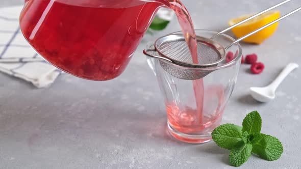 Woman hand is pouring raspberry tea in glass cup with sieve on a light background