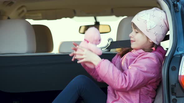 Pretty happy child girl playing with a pink toy teddy bear in a car trunk. alt