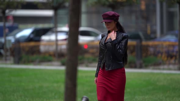 a Brunette in a Black Leather Jacket Red Skirt and a Burgundy Cap Walks Against the Background of a alt