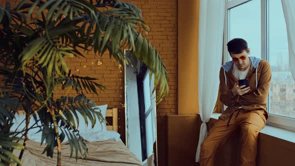 Young Man Sitting on Windowsill and Browsing Mobile Phone alt