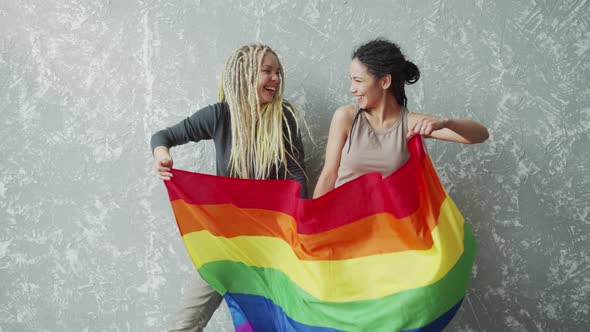 Positive Girlfriends Waving a LGBTQ Flag and Looking Into Each Other Eyes alt
