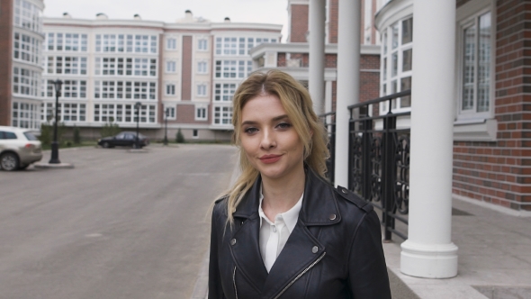 Young Woman Walking On The Street. Blonde Turns Flirting Gaze With The Viewer. Black Leather Jacket alt