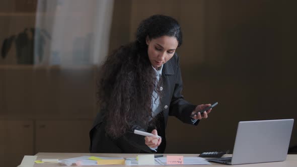 Focused Female Arabian Employee Designer Business Woman Standing at Office Desk Using Mobile Phone alt