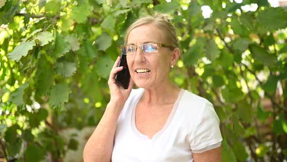 Elderly Senior Happy Woman in Prescription Eyeglasses Speaks Smartphone Outdoor