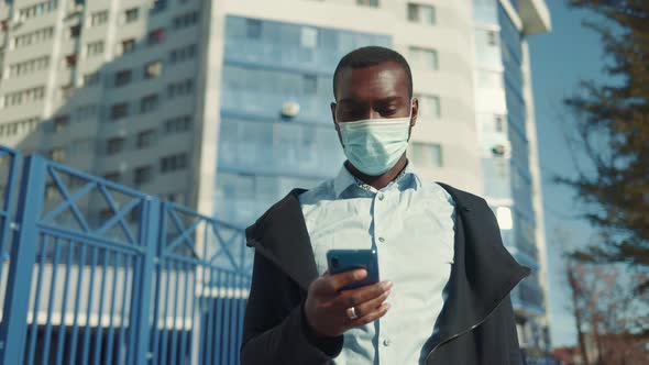 Portrait of African American Business Man in Mask, Smartphone in Hands. City Walk Down the Street alt