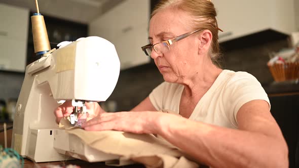 Confident Senior Elderly Woman Seamstress White Shirt and Glasses Sitting in Front of Sewing Machine