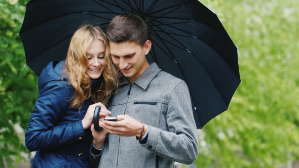 Young Couple Enjoys a Smartphone - Standing Under an Umbrella alt