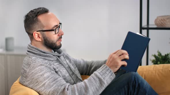 Confident Bearded Man in Glasses Reading Text Book Relaxing on Couch alt