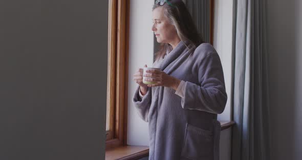 Woman drinking coffee while looking out of the window at home alt