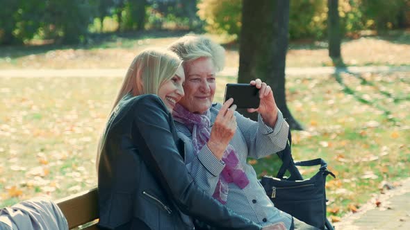 Zoom Shot of Young and Old Women Making Selfie on Smartphone in Autumn Time in a Beautiful Park alt