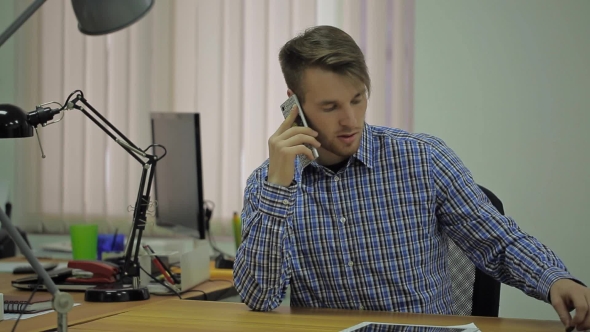 Young Man In a Blue Plaid Shirt Sitting At His Desk In The Office And Talking On a Smartphone alt