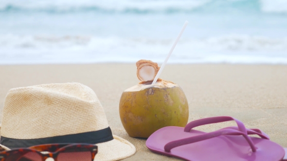 Coconut With Drinking Straw, Hat, Sunglasses And Beach Slippers On a Sand At The Sea