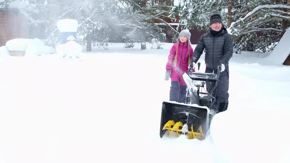 Man and his daughter in snowfall in yard remove snow with snowplow together.