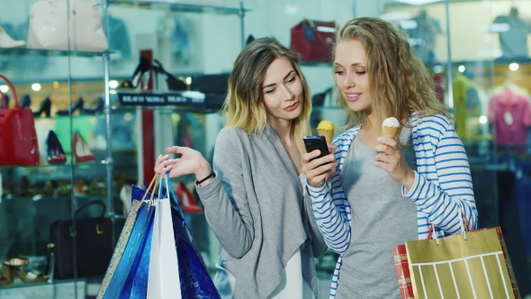 Two Young Women With Shopping Bags Dyal Go Ice-cream And Consider Something On The Phone. Smile, It alt