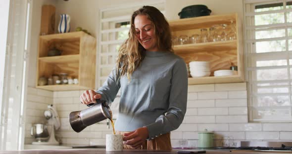 Happy caucasian woman standing in cottage kitchen pouring coffee from pot and smiling alt