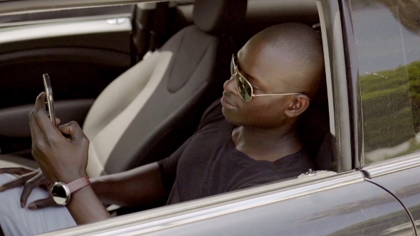 Handsome Black Man Seated In Car With Phone alt