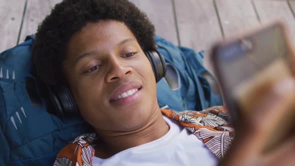Smiling african american man in city, lying on deck wearing headphones and using smartphone in park alt