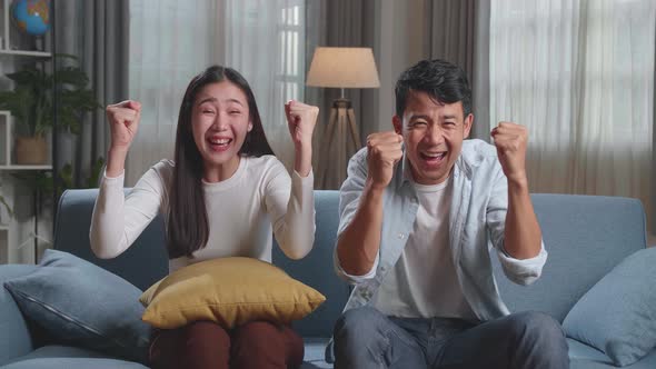 Excited Asian Couple Watching Tv Together Sitting On A Sofa In The Living Room At Home alt