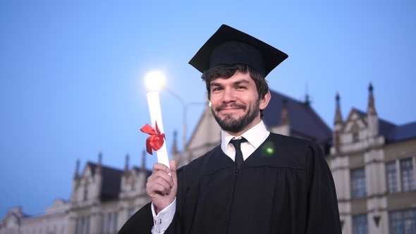 Guy in graduation outfit smiling and showing diploma to camera. alt