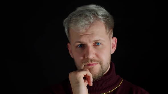 Cheerful Thinking Young Man Holding Hand on Chin Looking at Camera and Smiling on Black Background alt