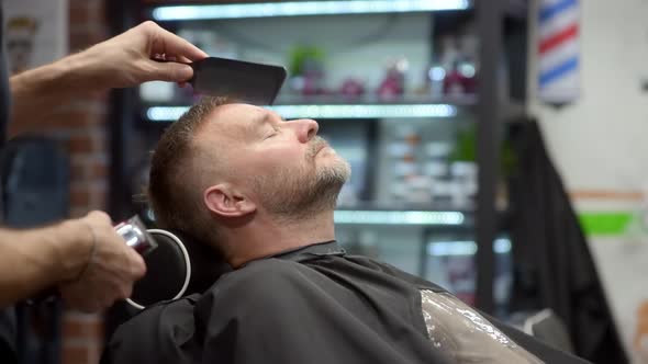 Barber master shaving handsome mature bearded man using electric shaver in salon