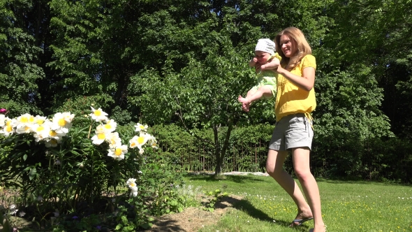 Happy Mom With Baby Girl Turn Around In Garden Near Peony Flowers.