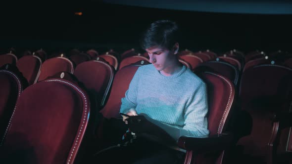 A lonely man in an empty theater is talking on the phone and watching the news. Looks at the phone.