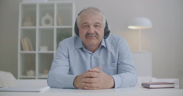 Middle-aged Man with Headphones on Head Is Nodding and Looking at Camera, Video Calling and Online alt