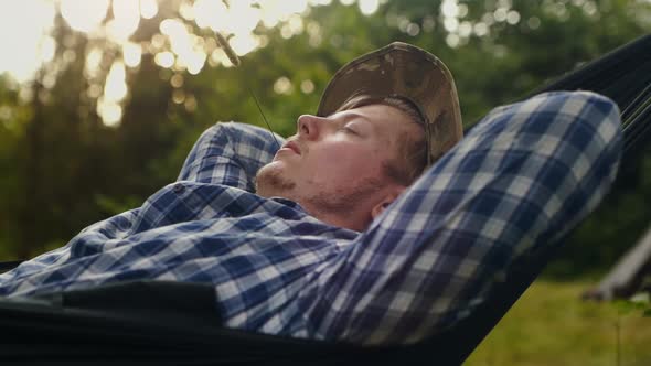 A Happy Man with a Hat and a Blade of Grass in His Mouth is Resting in a Hammock