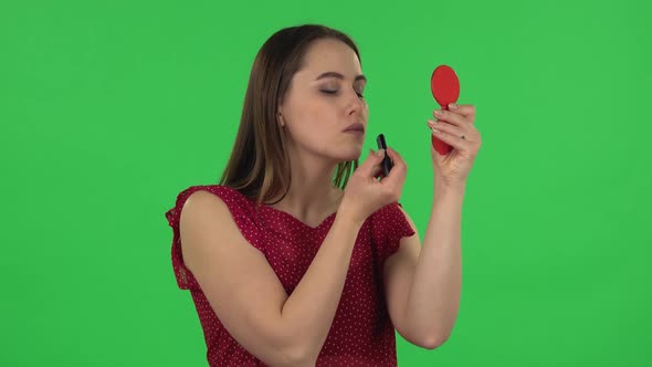 Portrait of Tender Girl in Red Dress Is Painting Her Lips Looking in Red Mirror. Green Screen alt