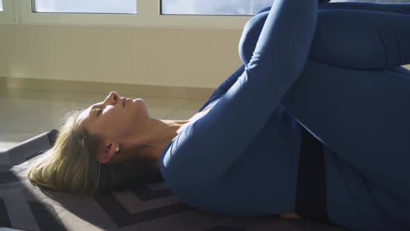 Woman in Blue Puts Knees To Chest on Floor in Sunny Gym