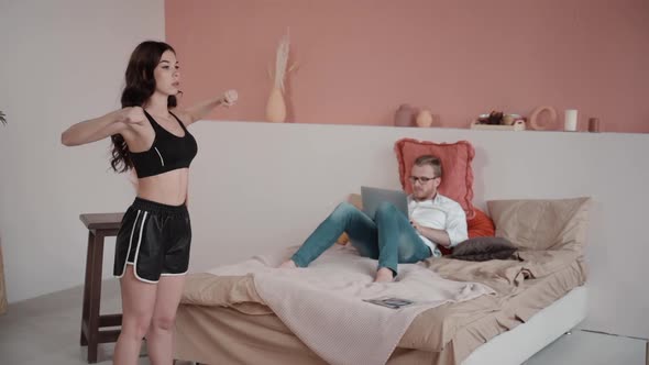 Young Woman Is Doing Exercises While Her Boyfriend Working with Laptop on Bed in Bedroom.