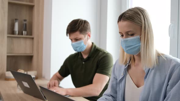 Caucasian Young Couple with Medicinal Mask on Face Are Working at the Laptop. alt