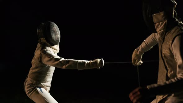 Two Fencers In Fencing Suits And Helmets Fencing In The Dark Outside. alt