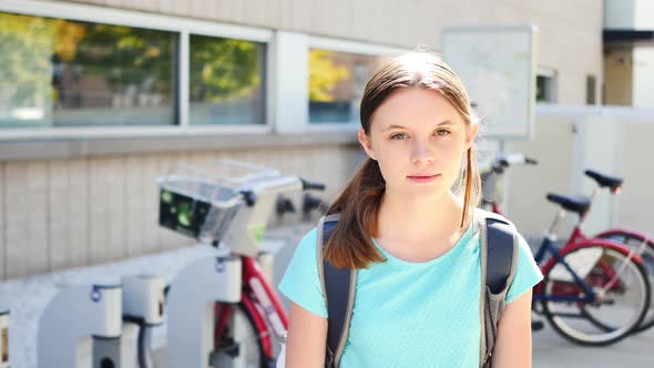 Portrait of inexpressive fifteen-year-old girl by city bikes who then smiles alt