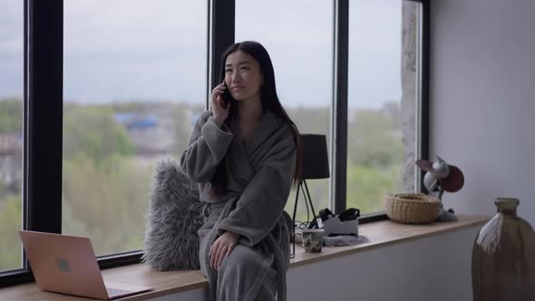 Cheerful Positive Asian Woman Talking on the Phone Sitting in Bathrobe on Windowsill in Home Office alt