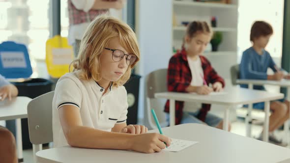 Redhead Pupil Wearing Glasses Sits at a Desk at School and Fills Out Tests and the Teacher in the alt