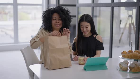 Young Smiling African American and Asian Women Unpacking Takeaway Food Sitting in Home Office at alt
