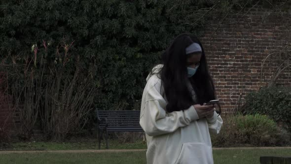 Female Teeanger Wearing Face Mask Checking Phone In Public Park During Lockdown. Locked off alt