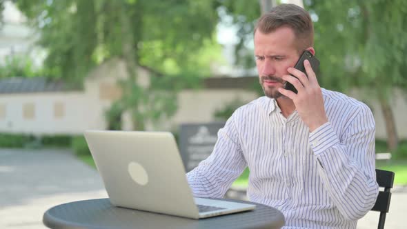 Angry Middle Aged Man Talking on Smartphone in Outdoor Cafe alt