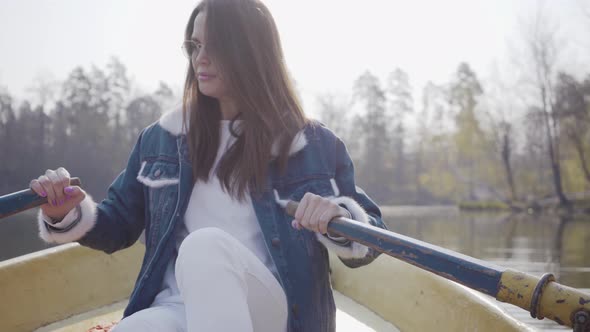 Pretty Glamour Woman in White Pants, Jeans Jacket and Sunglasses Paddles on the Yellow Boat on the alt