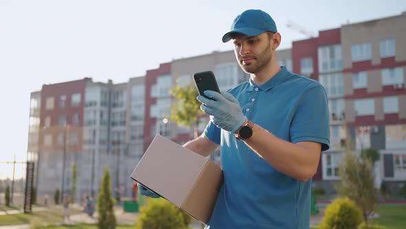 The Postman with Glasses Carries the Parcel and Looks at the Delivery Address Via Mobile Phone alt