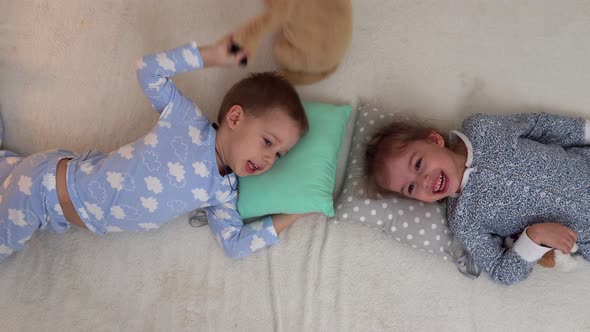 Two Smiling Preschool Toddler Children In Pajamas Playing With Teddy Bear on Bed