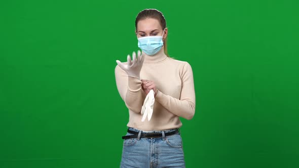Concentrated Young Woman in Coronavirus Face Mask Putting on Protective Gloves Posing on Green alt