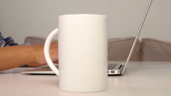 A Young Woman Drinks Coffee From a White Cup and Types on a Laptop Keyboard