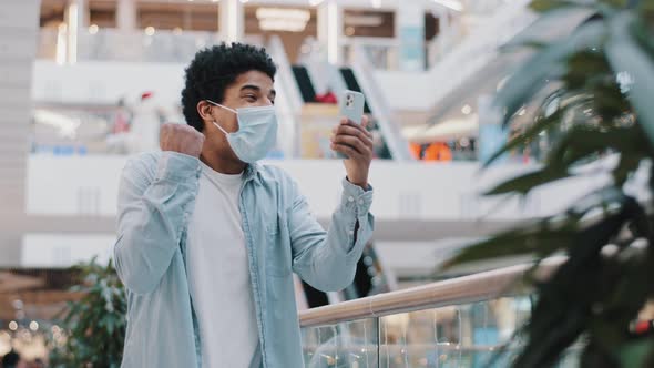 African Guy in Medical Face Protective Mask Stands in Mall Office Company Looking at Mobile Phone alt