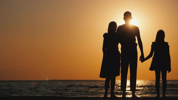 A Young Family with a Child Cuddles and Looks Forward to the Sunset Over the Sea alt