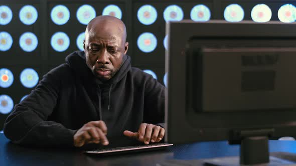 Bald Bearded Africanamerican Male Hacker in Black Hoodie Types on Keyboard